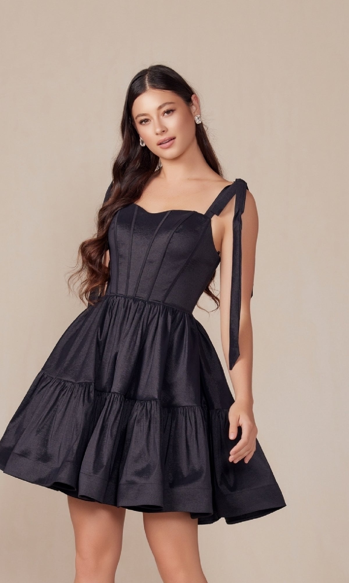 A woman with long, wavy hair faces the camera wearing the Shoulder-Tie Short Lace-Up A-Line Hoco Dress T869—a sleeveless black, knee-length A-line dress featuring a fitted bodice and flared tiered skirt.